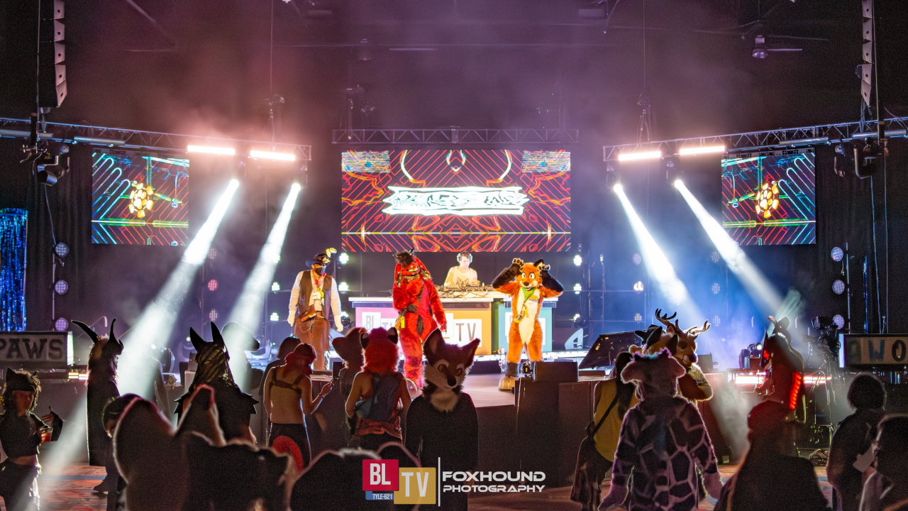 Main stage, illuminated with stage lights during a DJ set. There are fursuiters on stage and many people in the crowd below.