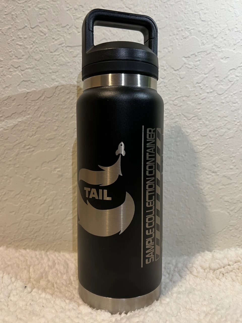 An image of the T.A.I.L. metal drink container, featuring a design of a rocket launching into space with a tail-shaped trail behind it. The side of the bottle says "Sample Collection Container".