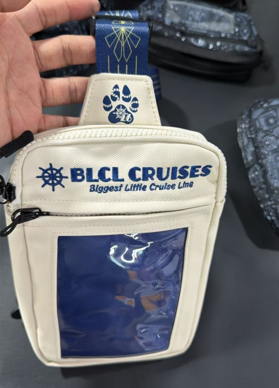 Cream canvas BLCL Cruises sling bag with clear front pocket, blue embroidery, and navy patterned strap.